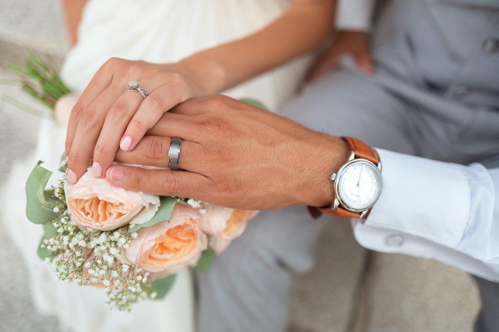 Close-up of a couple's joined hands resting on a small bouquet, wedding rings visible, the groom wearing a watch.