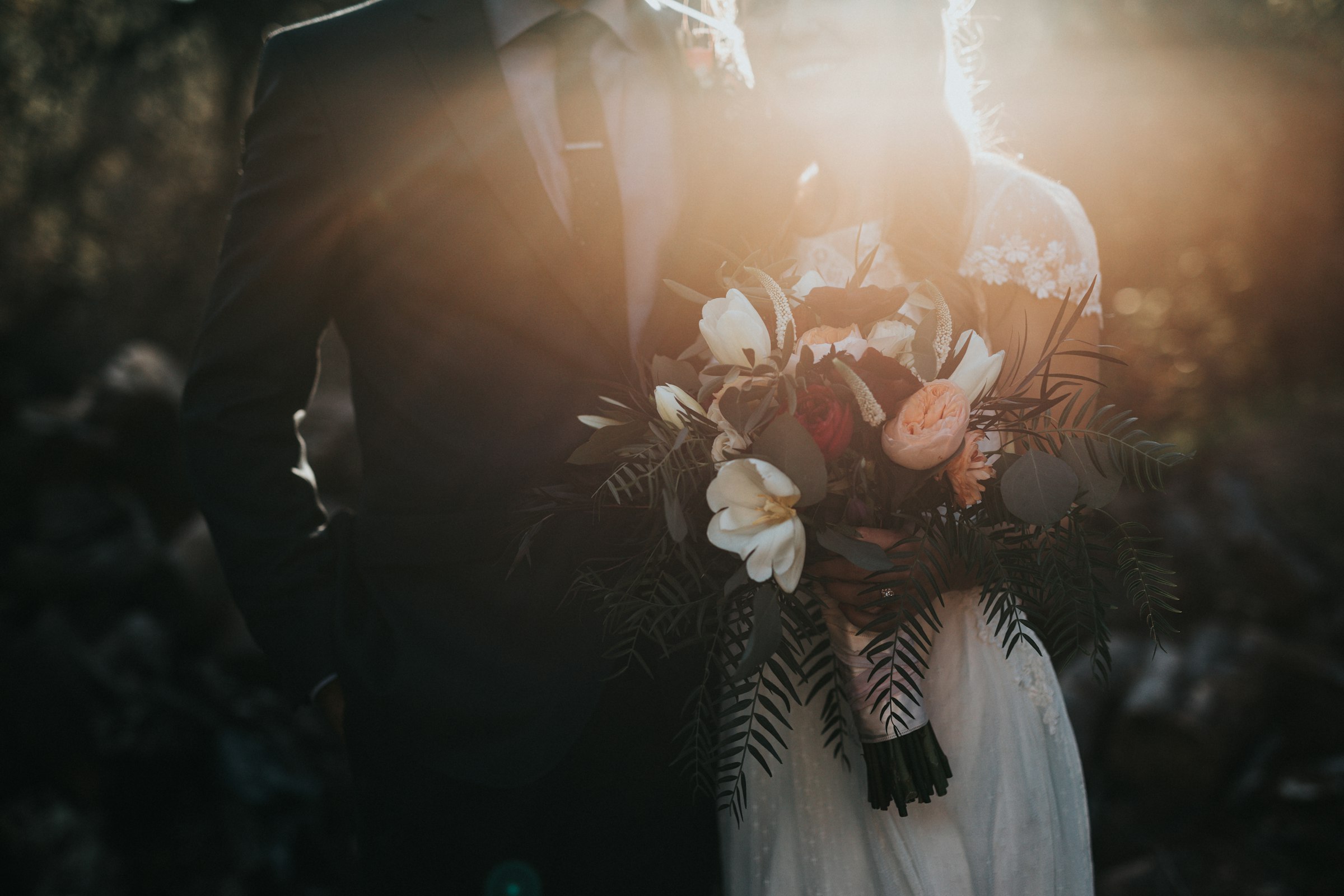 A couple standing close together outdoors at golden hour, the groom in a navy suit, the bride holding a bouquet, warm backlight around them.